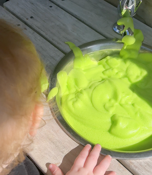 Red haired toddler peering into a bowl of green bubble foam in a mud kitchen